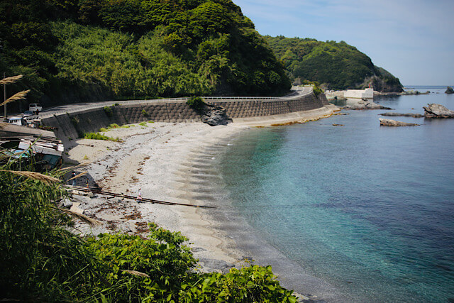 県道635号線から見下ろした海中線路の風景。透明度が高い青緑色の海へと、錆びたレールが砂浜から伸びている。海岸線に沿って走る県道と、緑豊かな山々、穏やかな水平線が広がる。