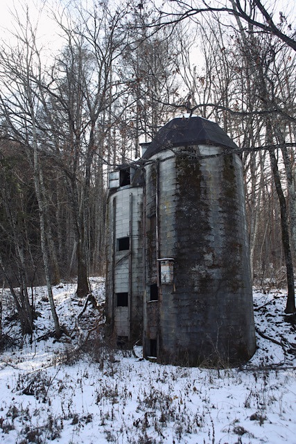 桑崎冬季分校の入り口付近に立つ、巨大なコンクリート製の円形サイロ。表面は苔むし、周囲の枯れ木や雪景色の中に孤立して建っている様子。