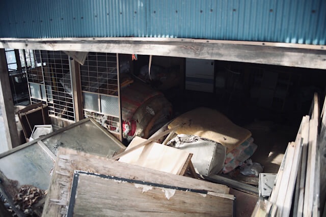 廃村となった桑崎集落の家屋内部。崩れ落ちた建材や窓枠の間に、かつて使用されていた布団や生活用品が散乱し、自然に還りつつある様子。