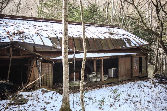 廃村・桑崎集落の奥に佇む大きな廃屋。剥がれかけた壁に「民宿」と書かれた看板が残り、屋根は歪み、周囲には雪が深く積もっている様子。