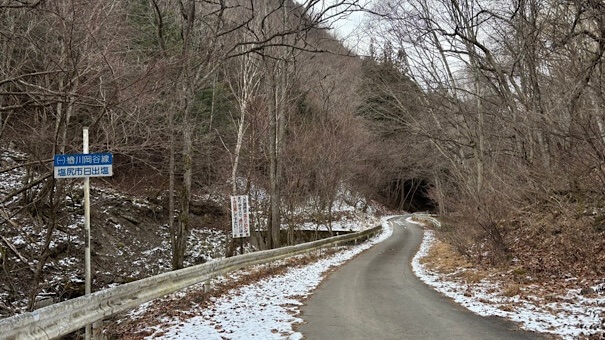 塩尻市日出塩付近の県道254号線(楢川岡谷線)。冬の枯れ木が左右から迫り、路肩に雪が積もった、車一台分ほどの道幅が続く山道の様子。