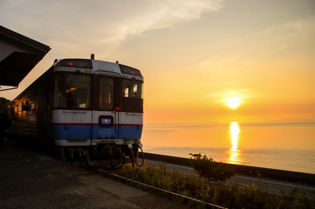 夕暮れ時の下灘駅。オレンジ色に輝く瀬戸内海を背景に、ホームに停車する八幡浜行きの列車。夕刻、黄金色の夕陽が海面と車体を照らしている情緒的な風景。