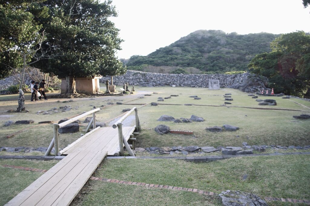 今帰仁城跡の主郭の風景。手前に木造の小さな橋があり、芝生の上には建物の土台となった石の遺構が点在している。背景にはなだらかな山と城壁が見え、観光客の姿はごくわずかで非常に空いている様子。