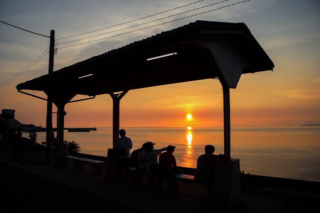 夕暮れ時の下灘駅ホーム。瀬戸内海に沈む夕陽を背に、ベンチに座って絶景を眺める人々のシルエット。太陽の光が人物の輪郭を黄金色に縁取り、レトロな駅の屋根が全体を額縁のように囲んでいる情緒的な風景。