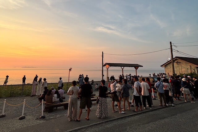 夕暮れ時の下灘駅。オレンジ色に染まる空と瀬戸内海を背景に、ホームや駅周辺に集まった大勢の観光客。多くの人がカメラを構えたり、撮影の順番を待ったりしている、お盆休みの混雑した様子。
