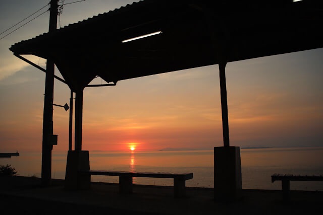 下灘駅のホームにあるレトロな屋根とベンチを黒いシルエットにし、その中央に瀬戸内海の水平線へ沈む夕陽を閉じ込めた額縁のような構図の写真。