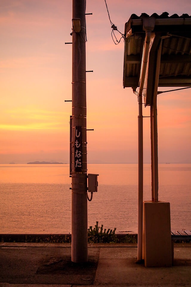 夕陽に染まる下灘駅のホーム。中央に「しもなだ」と書かれた駅名標の柱、右側にレトロな屋根の一部が見え、背景にはオレンジ色のグラデーションが広がる瀬戸内海が映っている。