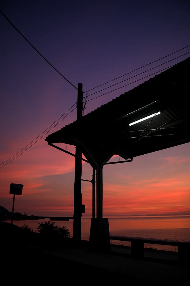 日没後の下灘駅ホーム。深い紫と燃えるようなオレンジ色のマジックアワーの空を背景に、駅の屋根とベンチが黒いシルエットとして浮かび上がっている。屋根の下には一本の蛍光灯が灯り、静かな瀬戸内海が広がっている。