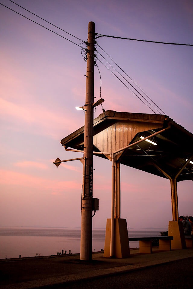 マジックアワーを迎えた下灘駅の風景。オレンジ色からピンク、そして紫へと変化する淡いグラデーションの空を背景に、「しもなだ」の文字が入った駅名標の柱と木造のホーム屋根が立っている。駅の明かりが灯り始め、静かな瀬戸内海が広がっている様子。