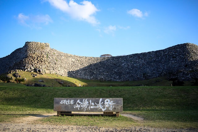 青空の下、緩やかな曲線を描いてそびえ立つ今帰仁城跡の巨大な石垣と、手前に置かれた「世界遺産 今帰仁城跡」の木製看板。