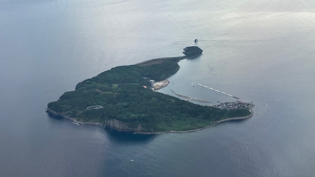 福岡空港へ向かう飛行機の窓から見下ろした相島の全景。深い緑に覆われた島と、白い防波堤がある港、穏やかな玄界灘が広がる空撮写真。
