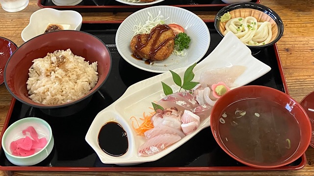 黒いお盆に載った豪華な刺身定食。魚の形をした皿に盛られた数種類の新鮮な刺身を中心に、炊き込みご飯、澄まし汁、ハート型のコロッケ、ミニうどん、冷奴、お漬物が並んでいる。