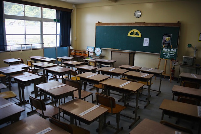むろと廃校水族館内の、当時のまま残された教室の風景。木の机と椅子が整然と並び、各机には「漁師川柳」が書かれた白い紙が貼られている。正面には大きな黒板と黄色の巨大分度器があり、そろばんや時計などの備品もそのまま展示されている様子。