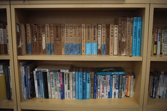 むろと廃校水族館の図書室にある木製の本棚。棚には『海洋生物学』『養殖学』『水産資源学』など、水産や海洋に関する漢字だらけの専門書が上下二段にぎっしりと並んでいる写真。