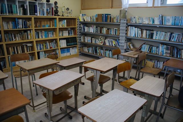 むろと廃校水族館の図書室。手前には昔懐かしい木の学習机と椅子が整然と並び、奥の壁際には専門書がぎっしり詰まった本棚が並んでいる。2025年8月に撮影された、学校の面影が強く残るノスタルジックな風景。