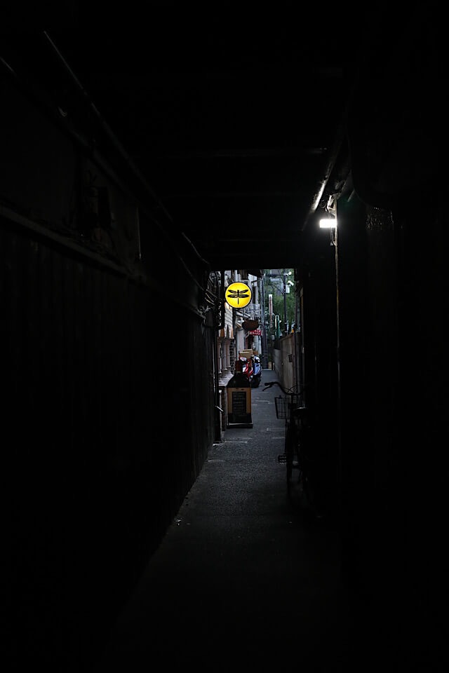 真っ暗で非常に細い路地の奥に、黄色い円形の看板（黒いトンボのマーク入り）が小さく光って見える風景。路地の壁に囲まれた狭さと、その先にある明るい出口のコントラストが印象的な一枚。