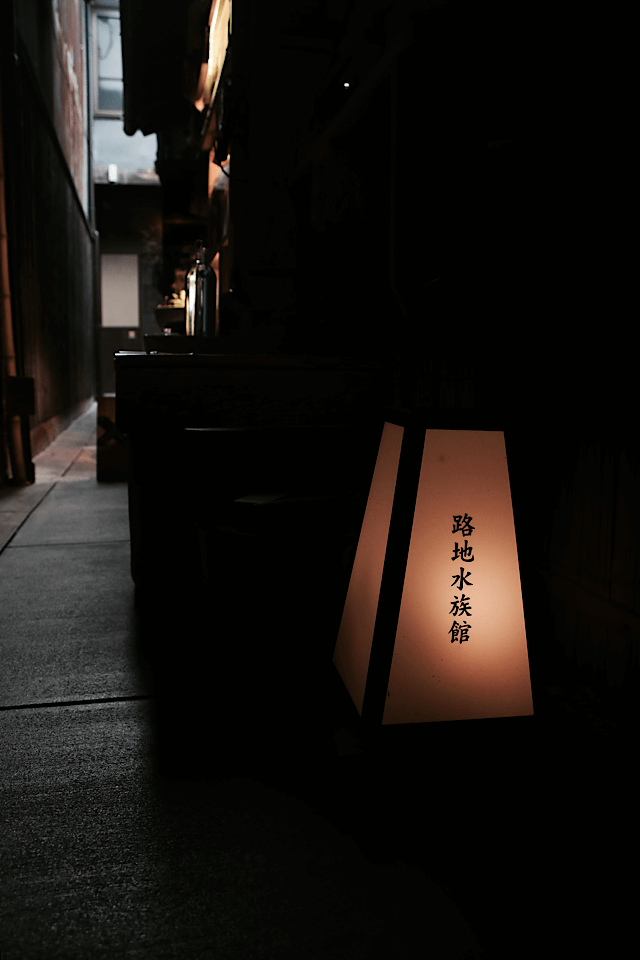 夜の京都・先斗町にある細い路地の入り口。手前に「路地水族館」と書かれた温かい光を放つ行灯があり、その奥に暗く静かな路地が続いている様子。