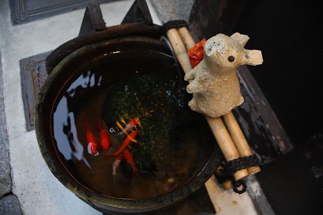 木製の桶の中を泳ぐ数匹の赤い金魚。桶の上には竹の棒が渡され、その上に小さなうさぎの石像がちょこんと座って金魚を見守っている。