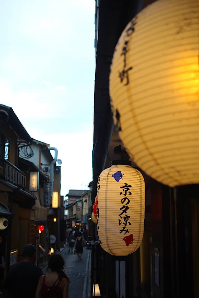 夕暮れ時の京都の通り。手前には「京の夕涼み」と書かれた大きな黄色い提灯が暖かく光り、その奥には伝統的な街並みの中を歩く人々の影が柔らかくぼやけて広がっている、情緒豊かな風景。