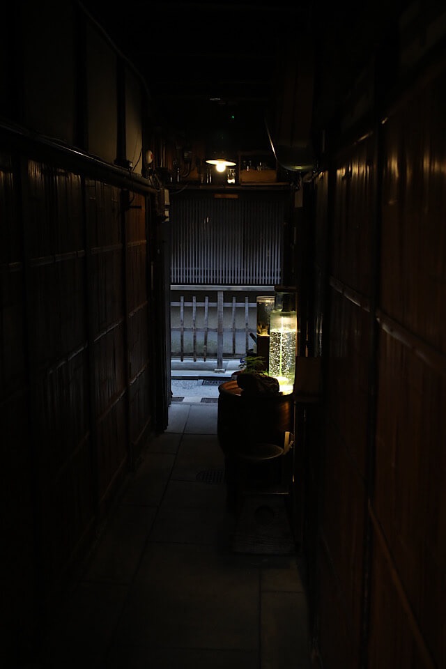 左右を古い木の壁に囲まれた、人ひとりがやっと通れるほど細くて暗い路地の風景。右側の足元には、幻想的に光る水槽やディスプレイが並び、奥へと続いている。