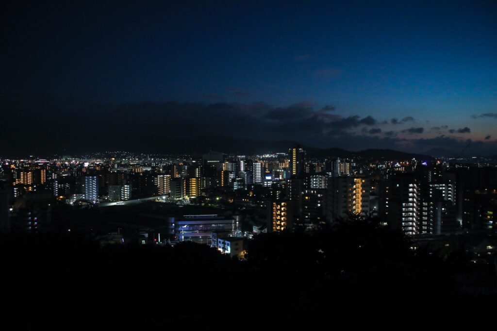 トワイライトタイムの福岡市街地の夜景。深い群青色の空の下、密集するビルの窓明かりや街灯が宝石のように散らばり、遠くに静かな山並みのシルエットが見える。
