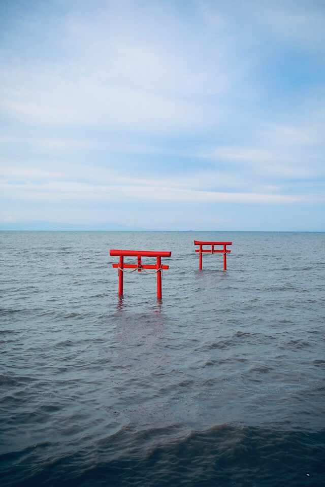 有明海の穏やかな波に包まれ、ジワジワと海に飲み込まれていく大魚神社の2基の赤い海中鳥居。青空と白い雲が広がる開放的な風景。