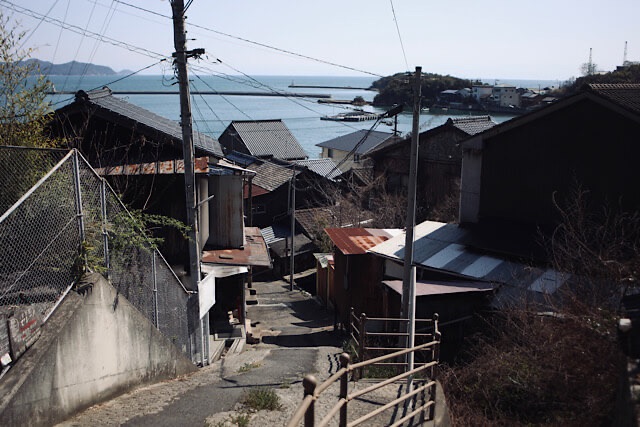 鞆の浦の坂道から瀬戸内海を見下ろす風景。古い瓦屋根の民家が並び、その先に穏やかな海と防波堤が見える、歴史を感じる渋い港町の日常。