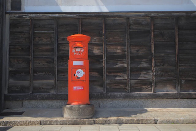 鞆の浦の歴史を感じさせる黒い木造の壁を背景に佇む、レトロな赤い丸型ポスト。港町の静かな日常に溶け込んだノスタルジックな風景。