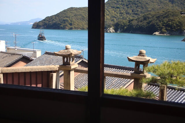 鞆の浦・福禅寺対潮楼の窓から眺める瀬戸内海。手前に並ぶ石灯籠と瓦屋根の向こうに、青い海を渡る「いろは丸」を模した渡船と、緑豊かな島々が見える風景。