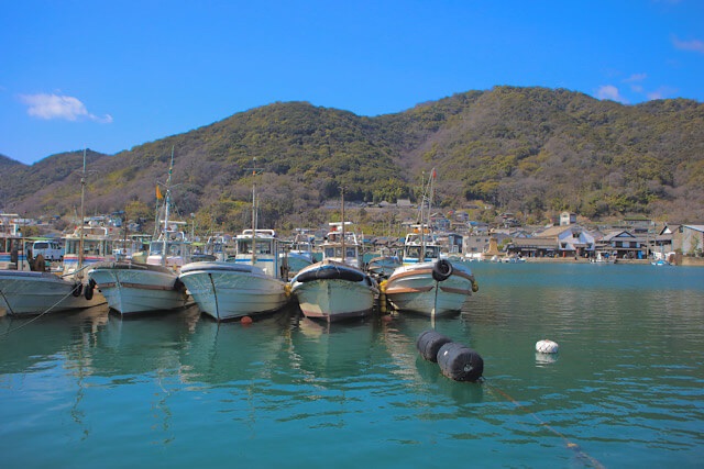 晴天の鞆の浦の港に並んで停泊する白い漁船。手前にはエメラルドグリーンの穏やかな海面が広がり、背景には春の陽光を浴びた緑豊かな山々と港町の家々が映し出されている。