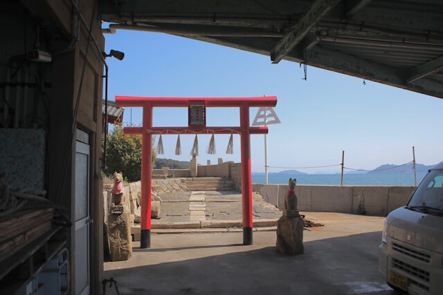 鞆の浦の港の片隅、建物に囲まれた場所に佇む鮮やかな赤い鳥居と2体の狐像（お稲荷さん）。背景には穏やかな青い瀬戸内海の水平線が広がっている。