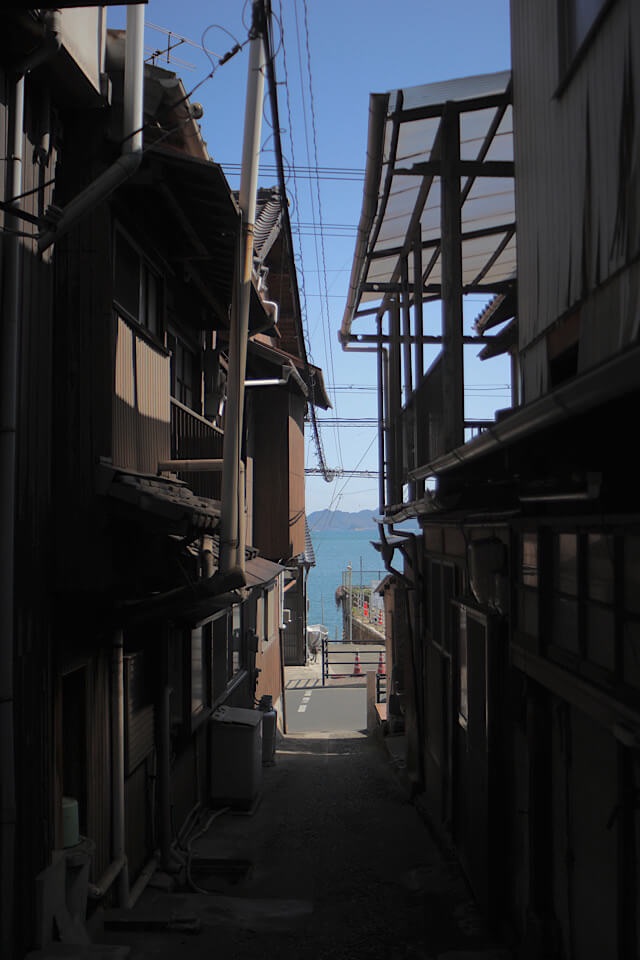 鞆の浦の古民家に挟まれた細く暗い路地裏。その建物の隙間から、鮮やかな青色をした瀬戸内海と遠くの島影が、額縁に収まった絵画のように見えている。
