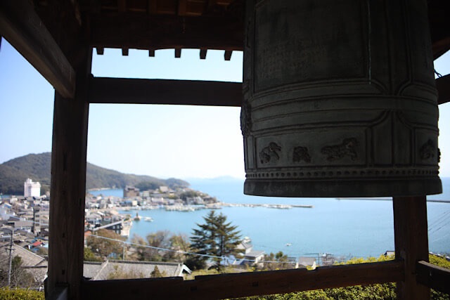 医王寺の大きな梵鐘と、その木枠越しに広がる鞆の浦の港町、穏やかな瀬戸内海、そして対岸の山々のパノラマ風景。