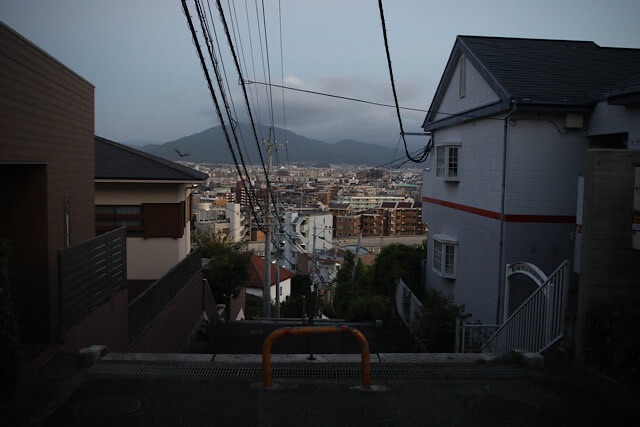 鷲尾愛宕神社への徒歩登山中、住宅街の狭い坂道の頂上から見下ろす、薄暗い街並みと遠くの山のパノラマ。手前にはオレンジ色のバリケードがある。街の灯りが少しずつ点き始めている。