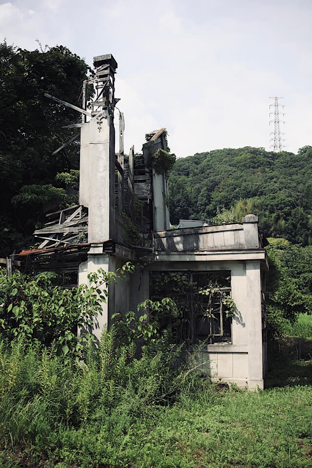 上を見上げた視点。旧仁堀郵便局の崩壊した屋根の隣に屹立する、コンクリート製(または石造り)の柱と、そのてっぺんに残る複雑な装飾。かつての「名建築のプライド」を感じさせる堂々とした姿は、崩壊した今でも気品を身に纏っている。