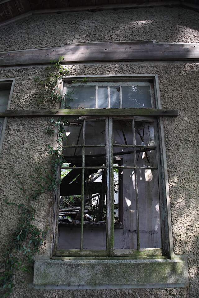 深い緑に囲まれた旧仁堀郵便局の、ガラスが割れ木枠だけが残る窓。外壁の蔦や窓の上の木の装飾が特徴。窓の奥には崩落した建物の内部が露出している。