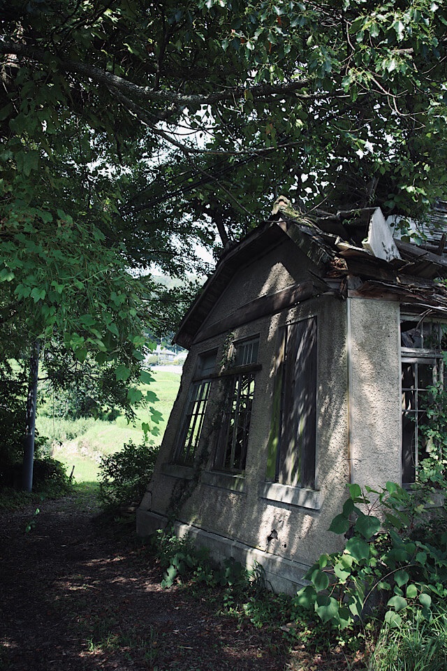 2025年8月撮影。深い緑に囲まれた、屋根が崩落し歪んだ窓が残る、末期的な半壊状態の旧仁堀郵便局の洋風外観。