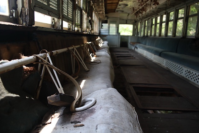 菊ヶ峠に保存されている片上鉄道キハ311形気動車の、老朽化により崩壊した車内。天井から剥がれ落ちた白い吊り革用の鉄の棒が、背もたれのクッションが剥がれたロングシートの上にドサッと横たわっている。鉄の棒には白い吊り革がぶら下がったまま、または座席の上に落ちている。床の一部が抜け、壊れた窓から夏の光が差し込んでいる。