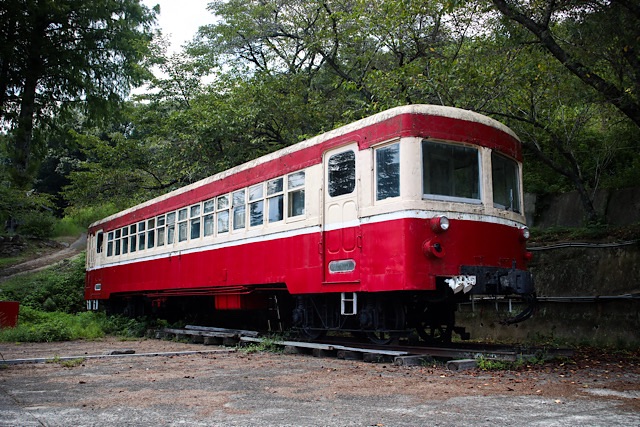 岡山県赤磐市の菊ヶ峠に静態保存されている、片上鉄道の赤とクリーム色のキハ311形気動車。夏の生い茂る木々を背景に、強い日差しを浴びて佇むノスタルジックな外観。