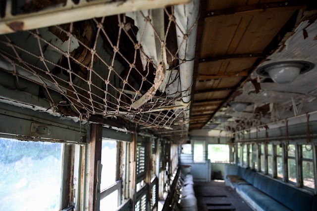 荒廃した片上鉄道キハ311の車内。前景には古く、一部が破れた網棚（網製の荷物棚）がクローズアップされ、天井板が剥がれ落ち、木製の構造、配線、古びた照明器具が露出している。時の経過を感じさせる光景。