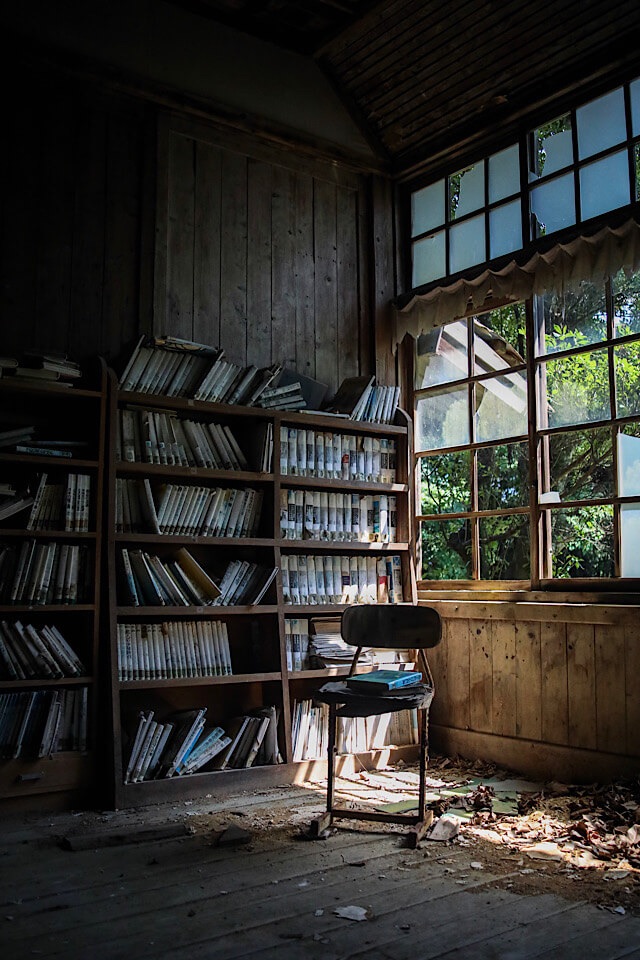 旧芳ノ沢小学校の図書室。木製の書棚には埃を被った大量の本が並んでいる。窓からは緑豊かな外の光が差し込み、その手前の木の床には枯葉や埃が散乱している。光の中に古びた児童用の椅子が1脚置かれ、その上には青い本が残されている。天井の木材も劣化が進んでいる。