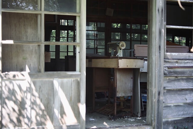 旧芳ノ沢小学校の木造校舎を、外の窓越しに覗いた光景。薄暗い室内には、重厚な木製の台と一体になった古い足踏みミシンが置かれている。色褪せた木の質感や窓枠の向こうに広がる静寂が、かつての家庭科室の記憶を留めている。