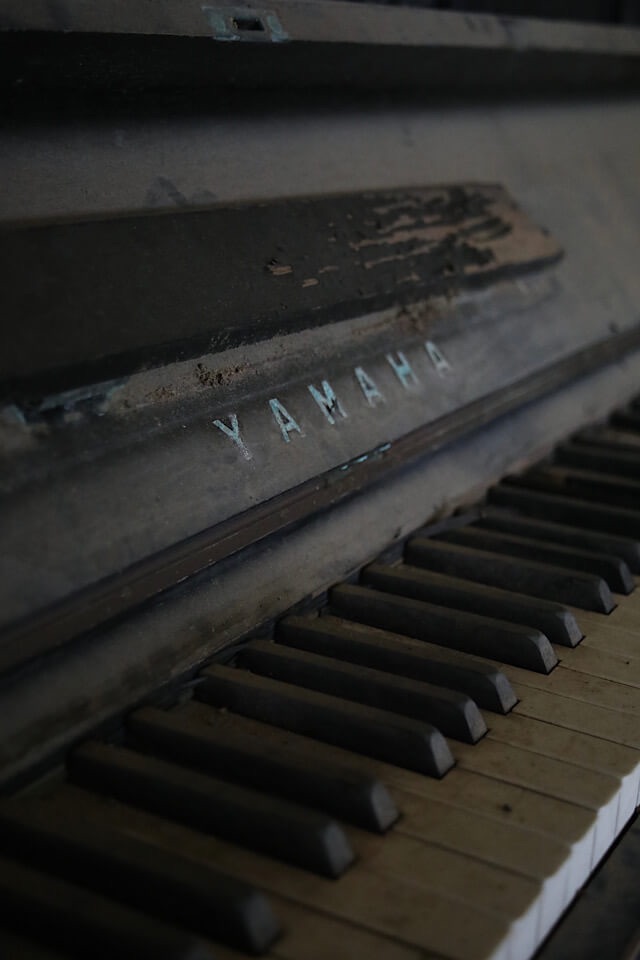 旧芳ノ沢小学校の音楽室。埃を被った古いアップライトピアノの鍵盤が、斜め上からのアングルで映し出されている。鍵盤の奥の木製の本体には「YAMAHA」のロゴが確認でき、塗装が剥がれ、長年の放置による劣化が目立つ。