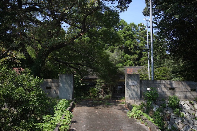 旧芳ノ沢小学校の校門。石垣のスロープの先に一対の灰色のコンクリート製門柱が立ち、その奥には生い茂る木々に囲まれた木造校舎の一部が見える。