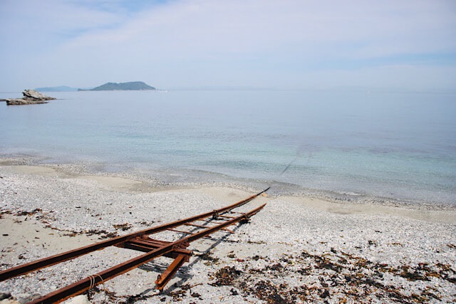 砂浜から海へと延びる錆びたレールは波打ち際で途切れており、その先の澄んだ青い海の中に線路が続いている。まるで『千と千尋の神隠し』の世界のような、遠景に島が見える風景。