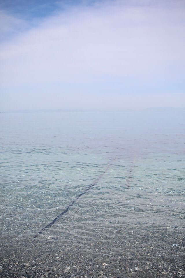 礫浜から、透明度が高い青い海へと伸びる錆びた海中線路。レールは途中で途切れており、その先が穏やかな海に沈んでいく様子。まるで『千と千尋の神隠し』の世界のような、遠景に霞んだ水平線が見える風景。