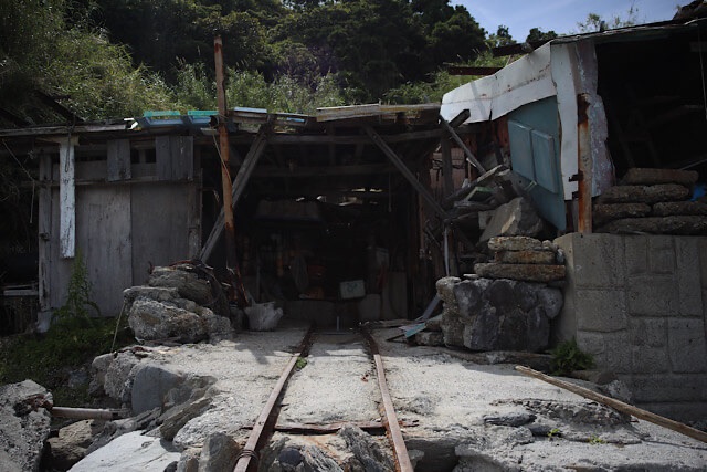 『海中線路』の起点に佇む古い建物。木とトタンでできた朽ちかけた小屋の中に、船を引き上げるための錆びた線路が続いている。背景には緑豊かな斜面が見える。