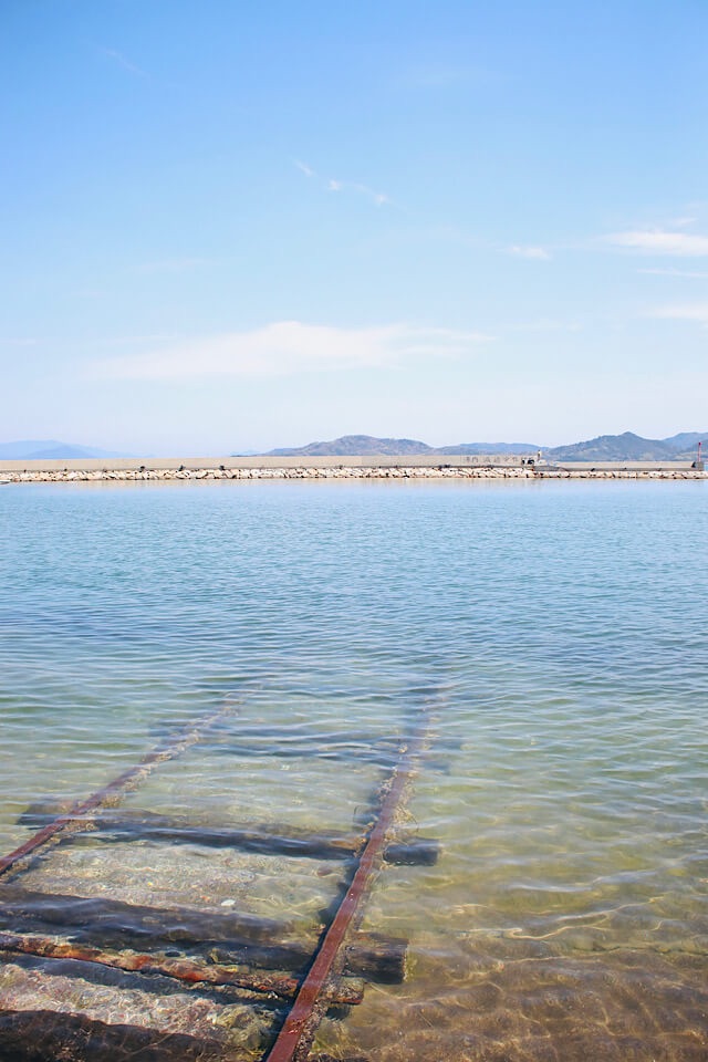 香川県房前漁港にある、澄んだ海へと真っ直ぐ伸びる錆びたレールと枕木の風景。穏やかな波と青空が広がる幻想的な海中線路。
