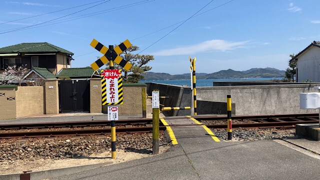 琴電志度線の線路にある、手動で押し下げるタイプの黄色と黒の縞模様の遮断桿が特徴的なレトロな踏切。黄色い「とまれみよ」の標識が立ち、踏切の先には青い海が広がっている。