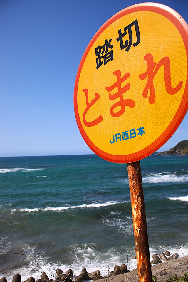 折居駅近くの海岸沿いに立つ、支柱が錆びた黄色い踏切標識「踏切 とまれ JR西日本」。背後には青空と、白波が立ち騒ぐ濃い青色の日本海が広がっている。